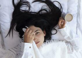 beauté femme veille en haut dans Matin et en buvant café dans lit. Jeune magnifique femme dans lit cache sa visage avec plaid et en portant tasse de café. mignonne Dame veille en haut. petit déjeuner dans une confortable chambre photo