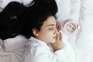 magnifique aux cheveux noirs femme en train de dormir dans confortable lit. le alarme l'horloge ensemble à sept un m Matin. femelle est Aller à réveiller en haut à sept heures de bonne heure Matin. Haut vue photo