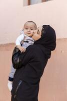 Jeune musulman femme dans hijab en portant sa 1 an bébé garçon, étreindre et embrasser. content musulman mère dans abaya Vêtements baiser et jouer avec sa peu enfant photo