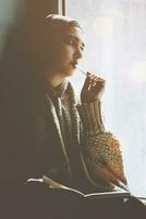Jeune musulman femme est assis sur le fenêtre seuil. moyen-orient écrivain femme séance pensivement près le fenêtre et l'écriture sa récit sur le bloc-notes. iranien femelle fabrication Remarques sur sa journal intime photo