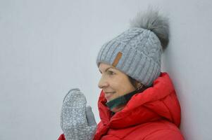 caucasien Jeune femme dans une rouge veste avec une capot, une gris tricoté chapeau et Mitaines sur une lumière Contexte. hiver portrait. photo