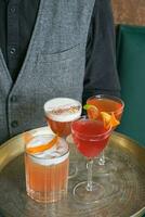 quatre brillant alcoolique des cocktails sur une plateau dans des lunettes dans le mains de une barman photo