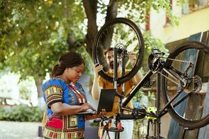 sportif copain et petite amie réparation vélo dégâts dans Accueil Cour en utilisant portable. actif et vibrant homme et noir femme sont explorant le l'Internet pour bicyclette entretien dehors. photo