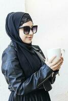 jolie milieu est fille dans traditionnel islamique vêtements, en buvant tasse de café. mignonne arabe femme en portant une tasse de chaud boisson dans mains photo