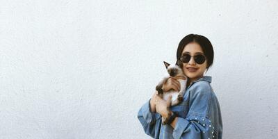 Jeune attrayant femme étreindre chatte chat dans mains. mignonne et glamour fille dans branché des lunettes de soleil posant avec sa Siamois chat photo