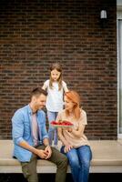 famille avec une mère, père et fille séance à l'extérieur sur pas de une de face porche de une brique maison et en mangeant des fraises photo