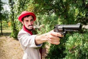 mime dans le apparence de une scélérat coups d'oeil en dehors de derrière une arbre et menace avec une pistolet. mal pitre avec une pistolet dans le parc. photo