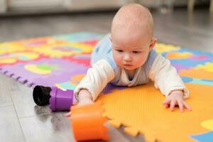 une bambin pièces sur le sol. le bébé est mensonge sur le sien abdomen sur tapis avec une pyramide. photo