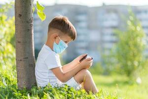 une enfant dans une médical masque est assis sur le herbe et regards dans le téléphone les dessins animés dans le été à le coucher du soleil. enfant avec une mobile téléphone dans le sien mains. la prévention contre coronavirus covid-19 pendant une pandémie photo