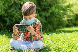 une enfant dans une médical masque est assis sur le herbe et regards dans le téléphone les dessins animés dans le été à le coucher du soleil. enfant avec une mobile téléphone dans le sien mains. la prévention contre coronavirus covid-19 pendant une pandémie photo