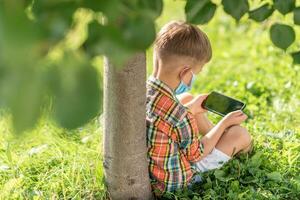 une enfant dans une médical masque est assis sur le herbe et regards dans le téléphone les dessins animés dans le été à le coucher du soleil. enfant avec une mobile téléphone dans le sien mains. la prévention contre coronavirus covid-19 pendant une pandémie photo