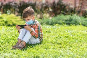 une enfant dans une médical masque est assis sur le herbe et regards dans le téléphone les dessins animés dans le été à le coucher du soleil. enfant avec une mobile téléphone dans le sien mains. la prévention contre coronavirus covid-19 pendant une pandémie photo