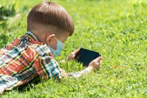 une enfant dans une médical masque mensonges sur le herbe et regards dans le téléphone les dessins animés dans le été à le coucher du soleil. enfant avec une mobile téléphone dans le sien mains. la prévention contre coronavirus covid-19 pendant une pandémie photo