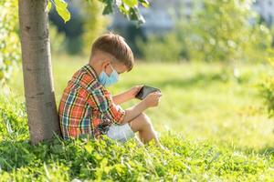 une enfant dans une médical masque est assis sur le herbe et regards dans le téléphone les dessins animés dans le été à le coucher du soleil. enfant avec une mobile téléphone dans le sien mains. la prévention contre coronavirus covid-19 pendant une pandémie photo