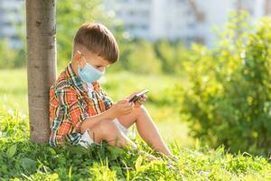 une enfant dans une médical masque est assis sur le herbe et regards dans le téléphone les dessins animés dans le été à le coucher du soleil. enfant avec une mobile téléphone dans le sien mains. la prévention contre coronavirus covid-19 pendant une pandémie photo