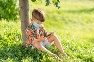 une enfant dans une médical masque est assis sur le herbe et regards dans le téléphone les dessins animés dans le été à le coucher du soleil. enfant avec une mobile téléphone dans le sien mains. la prévention contre coronavirus covid-19 pendant une pandémie photo