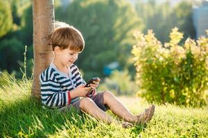 de bonne humeur garçon séance sur le herbe regards les dessins animés dans le téléphone dans le été à le coucher du soleil. mignonne bébé ayant amusement dans la nature photo