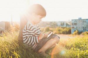 une magnifique enfant séance sur le herbe pièces sur le téléphone dans une Jeu dans le été à le coucher du soleil. garçon ayant amusement dans la nature photo