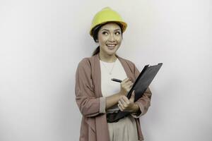 une réfléchi Jeune asiatique femme employé portant sécurité casque tandis que en portant une presse-papiers et à la recherche de côté à copie espace, isolé par blanc Contexte. photo