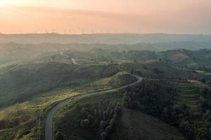 vue aérienne de l'autoroute incurvée sur la colline verte et de l'éolienne au sommet au coucher du soleil dans la campagne photo