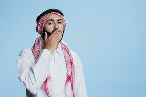 fatigué musulman homme habillé dans traditionnel vêtements bâillement et à la recherche à caméra avec somnolent expression. épuisé arabe la personne couvrant bouche avec main, fabrication ennuyé geste portrait photo