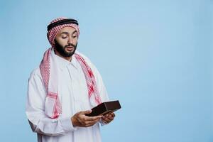 excité musulman homme habillé dans traditionnel vêtements en portant enveloppé cadeau boîte avec anticipation. surpris arabe la personne recevoir anniversaire cadeau, à la recherche à boite cadeau avec arc ruban photo