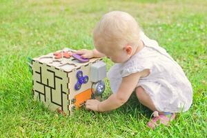 mignonne peu fille est en jouant avec tableau de bord en plein air sur vert herbe. éducatif jouet pour les tout-petits. fille ouvert porte à cube de planche. photo