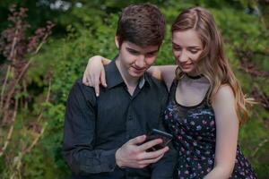 gars avec une petite amie fait du une selfie dans la nature. Jeune homme avec magnifique fille est à la recherche à le écran de noir téléphone. photo