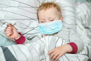 peu enfant dort dans lit dans protecteur médical masque. malade enfant dans blanc lit. enfant bambin un ans vieux avec grippe, grippe ou du froid protégé de virus, la pollution parmi les patients avec coronavirus photo