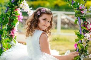 une magnifique fille dans un élégant rose robe est assis sur une balançoire décoré avec fleurs. enfant fête anniversaire 8 années. magnifique fleurs printemps thème balançoire dans jardin photo