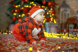 peu content fille de 6 mois rampe près une Nouveau année arbre sur Noël veille. enfant dans Père Noël claus costume près lumineux guirlandes. content Nouveau année 2020 concept photo