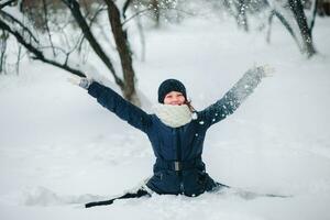 fille dans une noir veste et une noir chapeau est assis sur une chaîne dans le neige et jette neige. content enfant après une chute de neige. neige sur le des arbres. photo