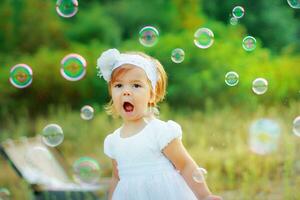 une peu fille dans une blanc robe et avec une fleur sur sa tête est content avec savon bulles. le enfant captures savon bulles. bébé en marchant dans le parc photo