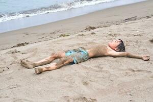 une garçon est mensonge dans le le sable après nager dans le mer. photo