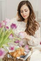 une cheveux bruns femme avec longue cheveux est ayant petit déjeuner à le table avec des croissants et une tasse de café. printemps portrait. photo