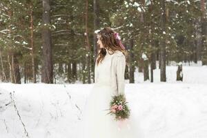 magnifique la mariée dans une blanc robe avec une bouquet dans une couvert de neige hiver forêt. portrait de le la mariée dans la nature photo