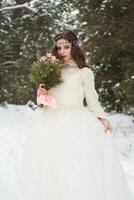 magnifique la mariée dans une blanc robe avec une bouquet dans une couvert de neige hiver forêt. portrait de le la mariée dans la nature photo