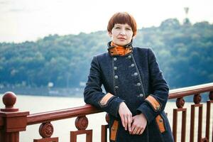 Jeune femme permanent sur une piéton pont dans kiev. le femme avec court cheveux penché sur le pont balustrade. femme sur le Contexte de Kiev paysage. photo