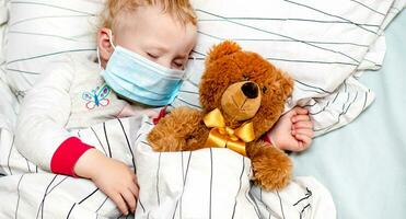 peu enfant dort dans lit dans protecteur médical masque avec une jouet ours. malade enfant dans blanc lit. enfant bambin un ans vieux avec grippe, grippe ou du froid protégé de virus, la pollution parmi les patients avec coronavirus photo