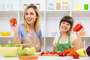 content mère et sa fille montrant tomate et poivre tandis que elles ou ils prendre plaisir fabrication en bonne santé repas ensemble à leur maison. photo