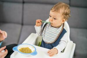 mignonne bébé garçon apprentissage à manger par lui-même tandis que séance dans une haute chaise photo