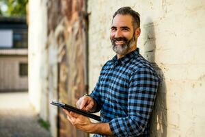 portrait de moderne homme d'affaire avec barbe en utilisant numérique tablette tandis que permanent dans de face de brique mur Extérieur photo