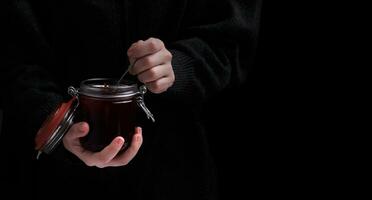 mains tenir une pot de fraise confiture et une cuillère sur une foncé Contexte. sélectif concentrer photo