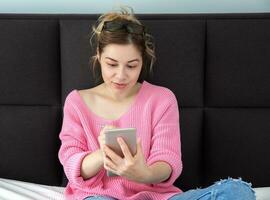 une Jeune fille avec blond cheveux est assis sur une canapé dans une rose chandail avec une téléphone, écrit ou dessine sur le écran avec une style photo