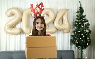 asiatique femme en portant une cadeau boîte avoir amusement célébrer Noël 2024. décorer et décorer le pièce avec une Noël arbre et 2024 des ballons. photo