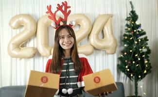 asiatique femme en portant une cadeau boîte avoir amusement célébrer Noël 2024. décorer et décorer le pièce avec une Noël arbre et 2024 des ballons. photo