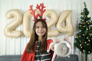 asiatique femme en portant une regarder avoir amusement célébrer Noël 2024. décorer et décorer le pièce avec une Noël arbre et 2024 des ballons. photo