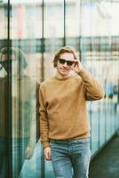 Extérieur portrait de Beau Jeune homme portant des lunettes de soleil et beige flou toison chandail, posant suivant à miroir mur sur ville rue Contexte photo