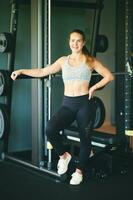 intérieur portrait de content Jeune en forme femme posant dans salle de sport, penché sur machine avec haltères, en bonne santé mode de vie photo