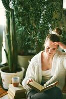 intérieur portrait de jolie Jeune fille relaxant à Accueil avec livres, femelle modèle séance sur sol, en train de lire vieux ancien livre, vert mis en pot les plantes sur Contexte photo